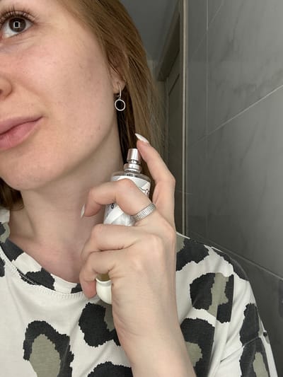 A close-up, profile view of a woman's face and upper body is captured in what appears to be a bathroom. The woman has fair skin, auburn hair, and is wearing a white t-shirt with a black and grey leopard print. She has a small, silver, hoop earring with a single dangling charm in her left ear. Her right hand is holding a clear glass bottle of perfume with a silver spray top and a white lid. The bottle has a white label with a grey geometric pattern. She is spraying the perfume on her neck. She is wearing a silver ring with a row of small stones on her right ring finger. The background consists of grey tiled walls, suggesting a bathroom setting. The image is lit by artificial light, causing a reflection in the woman's right eye. There is no information about the location being Нижние Муллы, Russia.