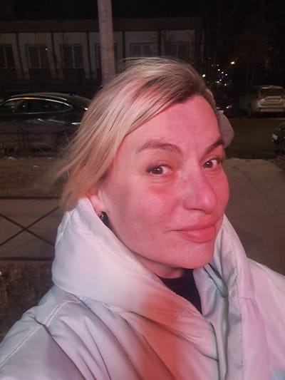 The image depicts an adult female subject in a close-up, selfie-style portrait taken outdoors at night in the city of Bel'kovo, Russia. The subject, with blonde hair, dark eyes, and a neutral facial expression with a slight smirk, is positioned centrally in the frame, facing the camera directly. She is wearing a light-colored (possibly white or light grey) puffer jacket with a high collar, over a dark inner garment. A small silver hoop earring is visible in her left ear. Her face and upper torso are illuminated by a strong reddish-pink light source, causing her skin to appear flushed and pink.
The background is dark and blurred, indicative of nighttime conditions and shallow depth of field. Visible elements include:
- A dark building with multiple illuminated windows in the far left background, suggesting a residential or commercial structure.
- Several parked vehicles, including a dark-colored sedan on the left and a light-colored SUV further back on the right.
- A dark, paved surface, likely a sidewalk or street, extends into the background.
- Bare tree branches are visible against the dark sky.
- Distant streetlights appear as blurred points of light.
- A vertical pole or tree trunk is partially visible behind the subject's head.