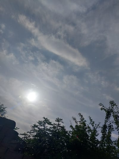 The image captures a sky with scattered clouds, a bright sun, and the silhouettes of trees and a wall. The sun is prominently visible in the upper left quadrant, casting a glare. Thin, wispy clouds streak across the sky, suggesting a partly cloudy day. The foreground is dominated by the dark silhouettes of trees with dense foliage, and a dark, textured wall on the left side of the frame. The scene appears to be outdoors, possibly in a rural or semi-urban setting given the presence of trees and a wall. The overall impression is a natural landscape under a bright sky, with the sun's intensity softening the details of the upper portion of the image.
