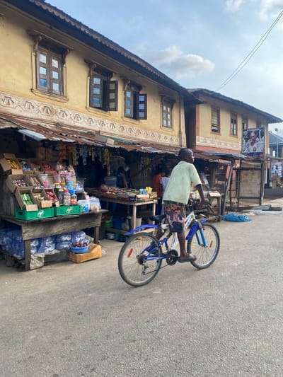 The image depicts a daytime street scene in Igarra, Nigeria. A two-story building with a pale yellow facade and a corrugated roof edge occupies the left side of the frame. Its upper floor features four wooden windows, two closed and two partially open, each with multiple glass panes. A decorative textured frieze runs horizontally beneath the windows on the upper floor.

Directly in front of this building is a market stall with a corrugated metal roof, supported by wooden poles. The stall displays various packaged goods, including snacks and candies, arranged in cardboard boxes and plastic containers on wooden tables. Green crates, some bearing a "7UP" logo, are repurposed as display units. Beneath the main display table, multiple bundles of clear plastic bottled water are stacked, and a blue basket filled with red onions rests on a wooden base. At least two other individuals are partially visible within the market stall on the left, one wearing an orange shirt.

In the mid-ground, an individual, identified as male, with dark skin, operates a blue and white mountain bicycle. He is wearing a pale green short-sleeved shirt, multi-colored shorts with orange, green, white, and blue patterns, and dark flip-flops. He is positioned standing over the bicycle frame, oriented with his back partially to the camera and gaze directed leftward along the street, indicative of movement. A red reflector is visible on the rear wheel of the bicycle.

Further down the street, on the right, a second two-story building with a similar architectural style is visible, featuring prominent reddish-brown trim on its corners and around its windows. A large banner depicting multiple figures and text is affixed to this building. A second market stall with a corrugated roof, less clearly detailed, is positioned further down the street on the right, fronting the second building. Two thin black electrical power lines traverse the upper portion of the light blue sky, which contains scattered white clouds. The street surface is paved asphalt, exhibiting signs of wear and minor irregularities.
