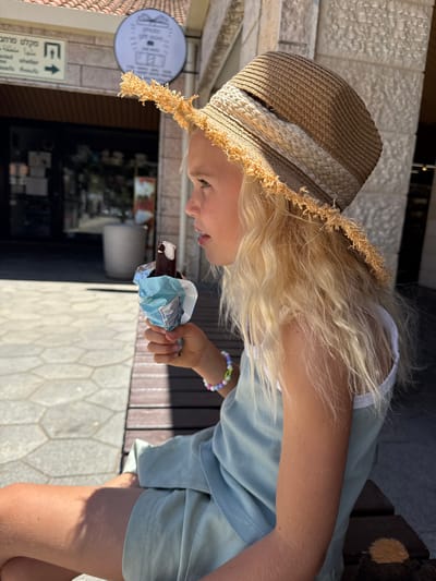 A young blonde girl is sitting on a wooden bench outside a shop in Kfar Vradim, Israel, eating an ice cream bar. She is wearing a straw hat with frayed edges and a light blue dress. The shop behind her has a sign with Hebrew writing and a circular logo. The pavement in front of the bench is made of hexagonal tiles. The lighting suggests it is a sunny day.