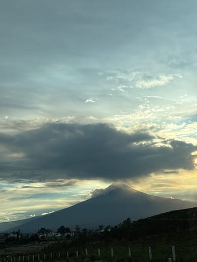 A wide, low-angle, outdoor, vertical shot shows the silhouette of a volcano against a sky filled with dramatic clouds. The volcano's peak is shrouded in wisps of white clouds, catching the golden light of the setting sun. The sky transitions from a pale blue at the top to a deeper grey and orange near the horizon, with patches of bright sunlight breaking through the clouds.
In the foreground, a dark, hilly landscape is visible, dotted with sparse trees. A small village with white buildings is nestled on the lower slopes of the volcano. A line of thin, pale fence posts runs horizontally across the bottom of the frame, adding a sense of depth to the scene. The location is identified as San Bernardino Chalchihuapan, Mexico.