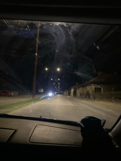 The image depicts a nighttime scene from the interior of a vehicle, looking through the windshield at a road. The road appears to be a rural or suburban street in Yasen, Bulgaria, lined with houses and streetlights. The houses are simple, with tiled roofs and dark facades. A red car is parked on the left side of the road. In the foreground, a dark, plush toy is visible on the dashboard. The windshield has streaks and smudges, some reflecting the streetlights and the dark sky. The overall impression is one of a quiet, dimly lit evening drive.