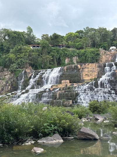 This image depicts the Pongour Waterfall in Tân Phú, Vietnam, on an overcast day. The waterfall cascades down a wide, multi-tiered rock formation, with streams of white water creating a frothy texture against the dark, layered stone. Lush green foliage covers the surrounding cliffs and the foreground, with dense bushes and trees thriving. In the distance, atop the cliffs, several buildings with red roofs are visible, nestled amongst the dense greenery. The foreground features a tranquil body of water reflecting the overcast sky and the surrounding vegetation. Several large, dark rocks are partially submerged in the water and scattered along the bank. The scene is natural and serene, showcasing the power and beauty of the waterfall and its lush, tropical environment.