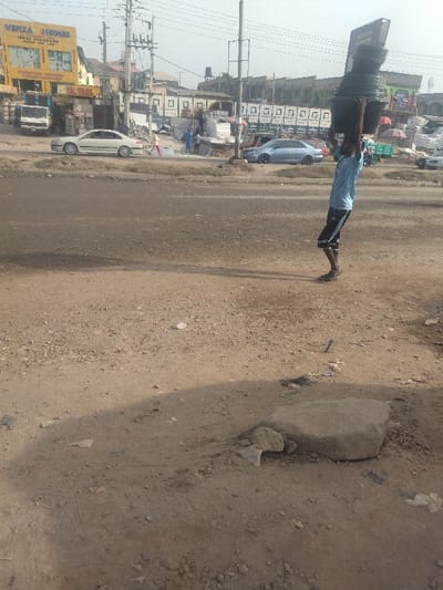 A man is walking down a dirt road in Way, Nigeria. He is carrying a large stack of black plastic containers on his head. There is a large rock in the middle of the road. There are several buildings in the background, including a yellow building with the words "VENZA HOMES" in large letters. The road is lined with power lines and poles. The man is walking in the direction of a blue car that is parked on the side of the road. The man is wearing a light blue shirt and black shorts. There is a small white car parked to the left of the road. A person in a red shirt can be seen walking away from the camera in the background. The ground is a mixture of dirt, rocks, and small debris. The sky is a light blue.