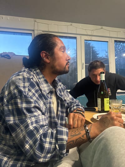 A man with dark hair pulled back in a bun, wearing a blue and white plaid shirt and light gray sweatpants, is seated at a wooden table. He has tattoos on his arms and hands. He is looking to his right, away from the camera. To his right, another man with short brown hair is wearing a black hoodie and looking towards the camera with his mouth open. A bottle of white wine and a glass of amber liquid are on the table between them. The room has large windows with a view of trees and the sky. String lights are visible on the ceiling. The setting appears to be a casual gathering in Ptichnoye, Russia.