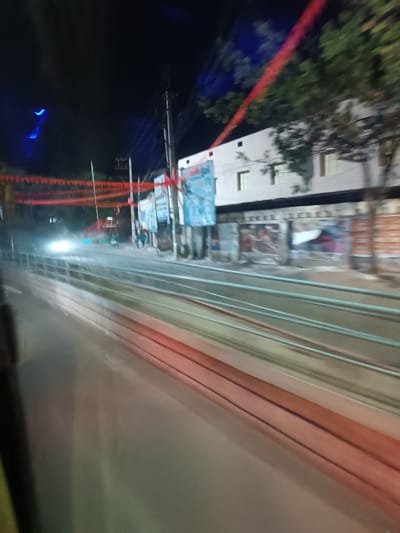 The image depicts a blurred, nighttime urban street scene in Anantapur, India, captured from a moving vehicle. The perspective is angled towards the right side of the road.

**Environment:**
The sky is dark, indicative of night. Red triangular bunting is strung horizontally overhead across the width of the street. Utility poles with multiple electrical wires are visible on the right, lining the road. Trees with green foliage are present behind and around the structures on the right.

**Objects and Structures:**
In the foreground, the paved road surface exhibits motion blur and reflections from light sources. A series of parallel horizontal railings, light in color, separate the roadway from the sidewalk and buildings on the right. Beyond these railings, a multi-story building, primarily white or light-colored, features several rectangular windows on its upper floors. The ground level of this building is covered with numerous large, faded, and partially torn posters or billboards, displaying various colors and indistinct graphics.

**Lighting and Activity:**
A prominent, intense white light source, likely the headlights of an oncoming vehicle, appears as a horizontal blur in the middle-left of the frame, suggesting vehicular traffic. Other distant, diffused lights are also visible. Blurred human figures are discernible on the sidewalk adjacent to the buildings on the right, indicating pedestrian presence. The motion blur across the entire image suggests the photograph was taken from a vehicle in motion.