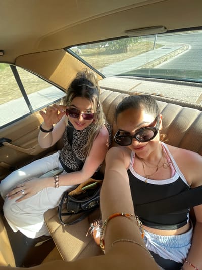 Two young women are inside a vehicle, possibly a car, taking a selfie. The vehicle appears to be moving, based on the blurred scenery visible through the windows. One woman is wearing a black tank top with a white trim and blue shorts, accessorized with multiple necklaces and bracelets. She is wearing large, dark sunglasses. The other woman is wearing a patterned blouse, white pants, and sunglasses. She has several bracelets on her wrist. The interior of the vehicle is beige. There are purses present. The exterior view suggests a road or highway setting. Based on the clues, it is not possible to pinpoint the exact location in El Puerto de Santa María.