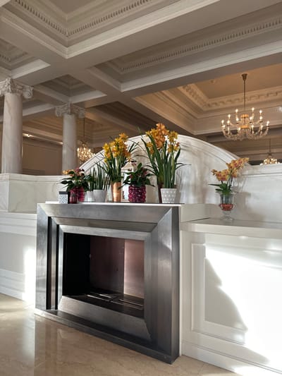 This image depicts a grand interior space within a building in Lausanne, Switzerland, characterized by neoclassical architectural elements. A modern, dark-finished geometric fireplace structure is centrally positioned in the foreground, featuring a recessed opening with a copper-toned back panel and a linear burner element. Above the fireplace, on a white marble mantelpiece, a varied arrangement of potted plants and decorative containers is displayed. These include red-flowered plants in textured clear glass pots, snake plants in textured ceramic pots, a copper-toned vase, and prominent arrangements of tall yellow-orange orchid-like flowers (Cymbidium) in white and copper pots. To the right, a smaller arrangement of similar yellow-orange orchids rests in an ornate clear glass vase on a lower white plinth.

The background reveals a spacious hall with high ceilings featuring elaborate white coffered molding and decorative cornices. Two large white fluted columns with ornate Corinthian capitals are visible on the left. Multiple crystal chandeliers, including a prominent multi-tiered fixture on the right and another further back on the left, illuminate the space. Strong directional light, likely natural, enters from the left, creating distinct shadows and bright highlights on the architectural surfaces and objects. The flooring is polished, light-colored tile or marble. No individuals are present, and no actions are occurring.