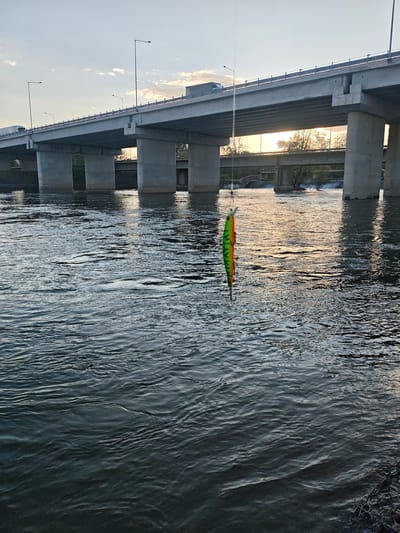 A fishing lure hangs suspended over the surface of a river. The lure is green, yellow, and orange. The river water is dark with ripples and reflections. In the background is a large concrete bridge with multiple supports and a road on top. The scene is illuminated by a setting sun visible between the bridge supports. The location is likely Strymoniko Municipal Unit, Greece, based on context. There are no people visible.