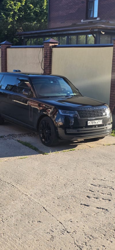 A black Range Rover is parked on a concrete surface in front of a light-colored stucco wall with a brick trim. The vehicle is facing the camera, with its front grille and headlights clearly visible. The license plate reads "P789KA977". Behind the wall, a building constructed of dark red brick is visible. Trees with green foliage are seen in the background. The scene appears to be a residential area in Русская Деревня-2 Кп, Russia, likely a private property given the wall and building. The concrete surface shows signs of wear and cracking. The lighting suggests it is daytime, with sunlight casting shadows. There are no people or other vehicles in the immediate vicinity.