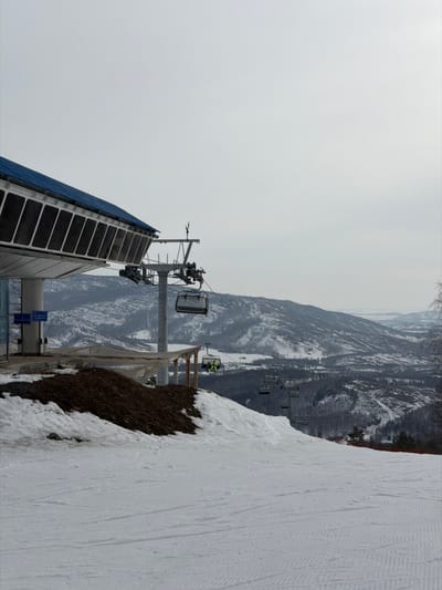 The image displays a winter ski resort scene in Novoabzakovo, Russia. In the upper left, a large, modern chairlift station features a light-colored metallic structure with a blue-domed roof and dark rectangular panels. Two blue informational signs are affixed to a support pillar of this station. Below the station, a groomed snow-covered ski slope extends into the foreground, showing visible ski tracks. A patch of exposed brown earth with dry vegetation borders the snow directly below the station structure. A chairlift cable originates from the station, supporting multiple grey chairlift carriers descending the slope. One chairlift carrier, positioned mid-frame, transports an individual wearing bright yellow outerwear, passing beneath a smaller, temporary shelter constructed with a white tarp roof and wooden supports. The background reveals extensive snow-covered mountains and hills, marked by dark tree lines, beneath a uniformly light grey, overcast sky.