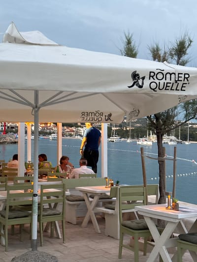 The image depicts an outdoor dining area with a view of the Adriatic Sea in Primošten, Croatia. A large white umbrella with the "Römer Quelle" logo provides shade for tables and chairs. Several people are seated at tables, engaged in conversation and dining. A waiter, wearing a blue shirt and black pants, stands in the background. The sea is calm, with several sailboats anchored in the distance. The shoreline is lined with buildings, and lush green hills form the backdrop. The overall atmosphere is one of a relaxed evening at a seaside restaurant.