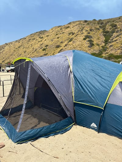 The image shows a blue and gray Ozark Trail tent pitched on a sandy surface. A grassy hill is visible in the background, and a blue sky is overhead. The tent appears to be in a campground location. The presence of the hill, sand, and tent indicates an outdoor recreational setting. The image was likely taken near a beach. Based on the vegetation type on the hill and the overall landscape features, the image could be taken in or near the city of Thousand Oaks in the United States.