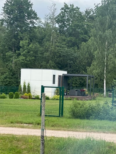 This media file captures an outdoor scene with a modern white building as the primary subject, situated amidst lush greenery and trees. The building appears to be a residential or commercial structure with a minimalist design, featuring a dark-trimmed window. Adjacent to the building is a dark-colored pergola, with outdoor seating visible underneath, suggesting an area for relaxation or entertainment.
The scene is bordered by a green metal fence, and beyond that, a gravel path runs alongside a well-maintained lawn. The background is dominated by dense foliage, indicating a natural or semi-rural setting. A single, thin birch tree with white bark stands out against the darker greens of other trees. The time of day appears to be daytime, with overcast or diffused lighting conditions, suggesting a cloudy or potentially rainy day.
There are no people visible in the image. The overall impression is one of a tranquil, modern property integrated into its natural surroundings. No text is discernible in the image. The location context provided is Astoņi, Latvia.