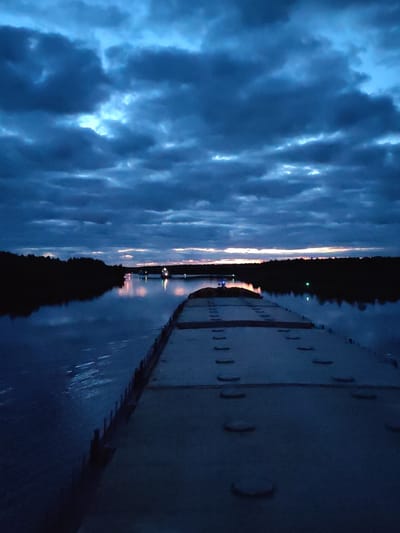 A nighttime shot showing a barge or large flat-topped vessel, possibly docked, on a body of water, presumably a river. The surface of the vessel has evenly spaced, circular protrusions. A dark line of trees and foliage is visible on both sides of the water, creating a shore. Light sources, possibly artificial, are seen in the distance, reflecting on the water's surface. The sky is filled with heavy, dark clouds, with slight illumination breaking through. The image is dark and appears to be taken at dusk or night. No people are visible. There is no verifiable information about the city of Uzhla, Russia.