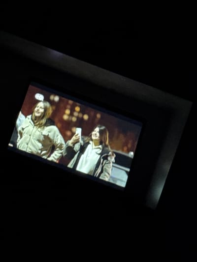 The image shows a close-up of a screen displaying two young women. The woman on the left is wearing a light-colored jacket and appears to be smiling. The woman on the right is holding a smartphone and is also smiling. The background on the screen is blurred and appears to be an urban setting at night, with bokeh lights. The screen is viewed at an angle, with the surrounding area being dark. The image is grainy and the resolution is not high. There is no discernible information within the image that indicates the location is Nasonovo, Russia.