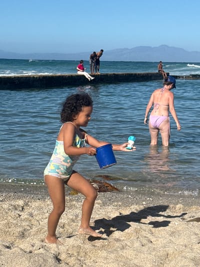 The image shows a beach scene in Muizenberg, South Africa. A young girl in a patterned bathing suit sits on the sand, holding a blue bucket and a toy. Behind her, an adult woman wearing a bikini is partially submerged in the water. In the background, a breakwater is visible with several people standing on it. The sky is clear and blue. The image is vertically oriented. The lighting suggests a sunny day.