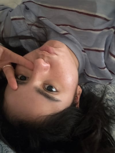 A close-up portrait shows a person, likely female, lying down and looking directly into the camera. The image is tilted approximately 45 degrees counter-clockwise relative to the subject's natural upright orientation. The subject has dark hair, fair skin with noticeable freckles across the nose and upper cheeks. Dark eyes are open, and dark eyebrows are present. The lips are full and slightly parted. The left hand is positioned with the index finger touching the skin below the left eye, and a thin silver ring is worn on the left ring finger. The subject is dressed in a light blue garment featuring thin horizontal stripes in white and dark red/maroon. The background appears to be a light-colored patterned fabric, suggesting an indoor environment. The scene is located in the city of Santiago, Mexico.