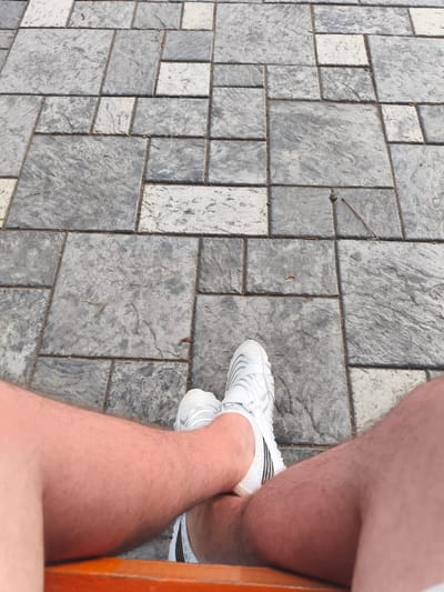 A first-person perspective shot shows a person's legs and feet resting on a concrete patio, likely outdoors. The patio is composed of rectangular and square concrete slabs of varying shades of gray, with some appearing lighter and whiter than others. There are some small brown debris pieces scattered between the slabs, possibly leaves or twigs. The person is wearing white athletic shoes with black stripes. The legs appear fair-skinned with visible hair. The lower portion of a reddish-orange bench is visible at the bottom of the frame. The location is identified as Bayet, Kyrgyzstan.