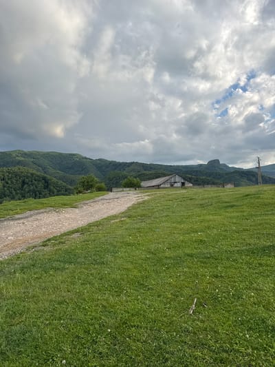The image depicts a rural, hilly landscape under a cloudy sky. In the foreground, a dirt road curves from the left towards the center of the frame, bordered by a grassy field. In the middle ground, a long, rustic wooden building with a sloped roof stands to the right of the road, with a low fence in front of it. Beyond the building, a distinct, rocky peak rises from the densely forested mountains that dominate the background. The sky is filled with dramatic, gray clouds, with some lighter patches suggesting breaks in the overcast. A wooden utility pole with wires is visible on the far right. The scene is devoid of any people or animals. The environment appears to be a natural, undeveloped area.