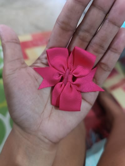 A close-up, overhead shot shows a hand holding a bright pink ribbon bow. The bow is intricately folded, with four pointed loops extending outwards from a central knot. The ribbon has a ribbed texture. The hand is open, with the bow resting on the palm. The background is blurred, with patches of color suggesting an indoor or outdoor setting. The lighting is bright, highlighting the details of the bow and the hand. The location of Kawit, Philippines, is not discernible from the image.