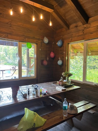 This is an indoor scene set in a rustic wooden cabin, likely located in the city of Bol'shaya Rechka, Russia. The image captures a dining area with a large wooden table in the foreground, covered with a clear plastic tablecloth. Scattered across the table are various items, including a red bag, several bottles, a metal object, a toothbrush, and a few other unidentifiable small objects. To the right of the table, a wooden bench is visible, with a plastic water bottle and a beige cap with "MIU MIU" logo placed on it.

The cabin's walls and ceiling are constructed from dark wood planks. A series of incandescent light bulbs hang from the ceiling, casting a warm glow. The room features large windows that look out onto a lush green forest with trees and foliage. Several colorful balloons (green, blue, and pink) are suspended by strings from the ceiling and walls, adding a festive touch to the otherwise simple decor. A vase with a bouquet of dried flowers sits on a tray on the table, next to a small chessboard. The floor is also made of dark wood planks. The overall impression is one of a casual, perhaps a vacation or a summer house setting, decorated for a small gathering. There are no people visible in the image.