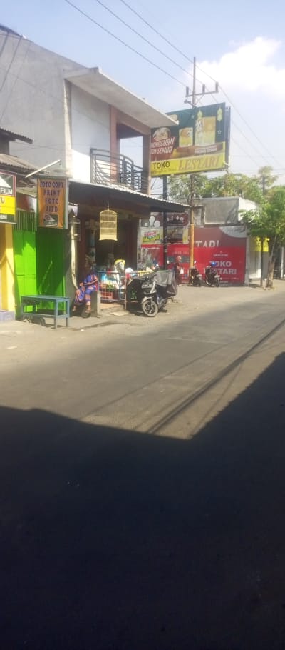 The image displays a daytime street scene in Kepuh Kiriman, Indonesia, characterized by commercial buildings, parked motor vehicles, and pedestrians under a clear sky.
The environment features a paved asphalt road in the foreground, partially covered by dark shadows cast from the left. Overhead utility lines are visible across the upper portion of the sky, supported by a utility pole in the midground. To the far right, green tree foliage is visible.
On the left, a two-story building has a ground-level storefront with a corrugated metal awning. The storefront displays a green metal gate and a yellow wall section. A white sign with red and blue text, advertising "FOTO COPY," "PRINT," "FOTO," and "JILD" (binding) with a Fujifilm logo, is affixed to the entrance. A light green wooden bench is positioned on the sidewalk in front of this shop. Inside the open shop entrance, a person wearing a blue patterned garment is seated on a low stool, facing outwards. Two traditional wooden bird cages are suspended from the awning structure. Various items, including a children's ride, are visible inside the shop. A black motor scooter with a large rear delivery box, license plate M 5447 QAX, is parked in front of this shop.
Further to the right, a multi-story building features a prominent red-painted ground floor facade marked "TOKO LESTARI" and "DANDANG." A billboard above this building advertises "HONGGUAN TUK SEMUA" and "TOKO LESTARI," displaying images of food products. Multiple motor scooters are parked in front of Toko Lestari. At least two individuals are visible near the entrance of Toko Lestari; one individual wearing a headscarf is partially obscured. Another sign advertising "Yakult" is visible near the Toko Lestari entrance.