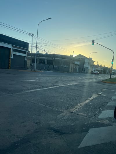 The image depicts a street view in Gerli, Argentina, during the daytime. A building with a partially visible facade stands alongside the road. The sky appears bright, likely due to sunlight. Overhead, power lines are visible, intersecting the view of buildings and other street infrastructure. The asphalt road shows signs of wear and tear, with cracks and patches. A traffic light is situated at an intersection, indicating a green light. The focus is on the urban landscape, with buildings and streets.