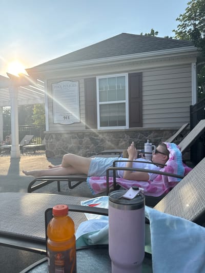 A young person with blonde hair, wearing sunglasses and a blue and white striped skirt, lies on a lounge chair by a pool. They are looking at their phone, with a pink and blue towel draped over their head and shoulders. The sun is setting, casting a warm glow on the scene. A building with "POOL POLICIES" signage is visible in the background. In the foreground, a bottle of orange liquid and a pink tumbler sit on a table. The location is the pool area of a community in Allen Township, United States of America.