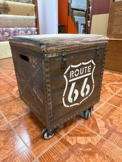 The image shows a close-up, eye-level view of a vintage-style wooden trunk with a cushioned top, resting on casters. The trunk is dark brown with distressed wood grain and metallic accents, including rivets and latches. Prominently displayed on the front is a white stencil of the "Route 66" shield logo. There is also text stenciled on the side of the trunk, including "3-85" and "HIGH OPERATION 0025-TOM-A45", along with other illegible markings. The trunk sits on a floor with terracotta-colored tiles that have a wood-grain pattern. In the background, out of focus, are rolls of patterned wallpaper, wooden panels, and shelves containing various items, suggesting an indoor storage or workshop environment. The scene is well-lit, with natural light entering from an unseen source, casting soft shadows. There are no people visible in the image. The location is not identifiable as Maloyaroslavets, Russia, or any other specific city based on the visual information provided.