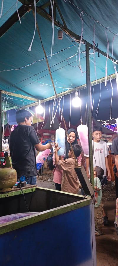 The image depicts an outdoor market scene, likely at night, given the artificial lighting. A stall is set up under a teal-colored tarp, supported by wooden beams. Strings with plastic streamers hang from the structure, along with bare light bulbs that illuminate the area.
In the foreground, a man wearing a black t-shirt and blue jeans stands behind a counter, which has a green-painted wooden frame and a blue lower section. A green gas tank is visible on the left side of the counter, connected to some electrical components.
Interacting at the stall are a woman wearing a black hijab and a pink top, and two boys. One boy, in a brown shirt, is reaching up towards a large, translucent blue bag of cotton candy. The woman is looking at the boy and appears to be smiling. Another boy, in a light green t-shirt and khaki shorts, is standing to the right, looking up. A young man in a white t-shirt with red lettering and a young child are also visible further back, as are other people in the background, suggesting a crowded event. The ground is covered in loose gravel and debris.
The scene captures a moment of commerce and leisure, likely during a local festival or market day in Sinar Semendo, Indonesia, as suggested by the context. The cotton candy, a common fairground treat, and the general atmosphere point to a community gathering.