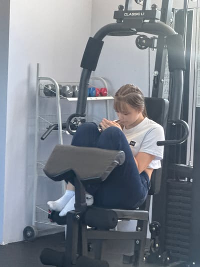 The image shows a young woman using a leg extension machine in a gym. She is wearing a white t-shirt and dark jeans, and is seated on the machine with her legs positioned under the padded lever. A rack of dumbbells is visible in the background. The machine has "CLASSIC L1" printed on it. The location is Obruchishte, Bulgaria.