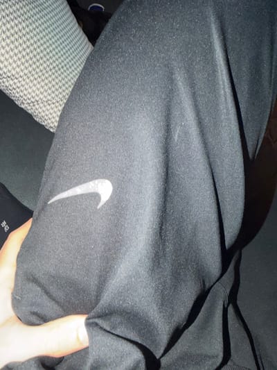 The scene, situated in Tuttlingen, Germany, depicts a close-up, downward-angled view focusing on an individual's lower leg or thigh. The primary subject is a section of black athletic apparel, identified as Nike by a silver-white "swoosh" logo positioned on the fabric's left side. The garment material shows a slight textured appearance and minor pilling. A light-skinned human hand, with visible knuckles and fingernails, is gripping or resting on the black fabric in the lower-left portion of the frame. In the upper-left background, a segment of a light-colored object with a small, repetitive geometric pattern, consistent with upholstered seating or a cushion, is visible. A sliver of black fabric with white numerical markings ("42-42"), indicative of a sock or another article of clothing, is discernible beneath the hand. The lighting appears to be from an internal source, casting shadows and highlights across the fabric's contours.