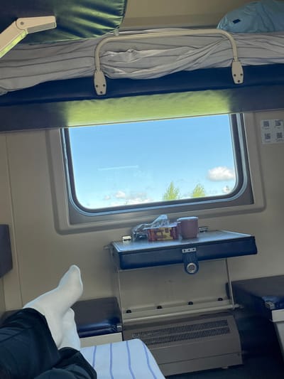 The image captures an interior view from a bunk bed in a train compartment, looking out a window towards a bright blue sky with scattered clouds and distant green trees. In the foreground, a person's white-socked feet and lower legs, clad in dark pants, are visible, resting on the lower bunk. A small fold-down table attached to the wall is positioned below the window, holding a box of tea, a pink mug, and some plastic-wrapped items. The compartment features two bunks, with bedding visible on both. The overall impression is that of a traveler relaxing during a train journey. This scene is located in a train compartment, not a specific location in the city of Нижняя Корса, Russia.