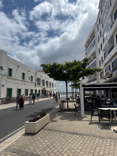 The image depicts a street scene in Arrecife, Spain. The street runs between two buildings; one tall and modern, and the other a traditional two-story building with white walls and green windows. The street is paved with asphalt, and the sidewalk is made of light gray paving stones. Planter boxes line the street. People are walking along the street. A tree provides shade near an outdoor cafe with customers seated at tables. The sky is cloudy with patches of blue.