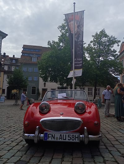 In Meersburg, Germany, a vintage red convertible car is parked on a cobblestone street. The car, identified by its license plate "FN AU 58H", is the primary subject, positioned centrally in the foreground. Its headlights are large and round, resembling eyes, and the grille forms a smiling mouth, giving the car a cheerful appearance. Behind the car, a tall banner hangs from a pole, featuring a portrait of a man and text that appears to be in German, possibly indicating a historical event or exhibition.
Several people are visible in the background. To the left, a man in a light blue shirt and shorts is pushing a bicycle. Further back, groups of people are walking and standing, observing the scene. The environment consists of historic buildings with traditional German architecture, including pitched tiled roofs and multi-story facades. Green trees provide a natural element against the urban backdrop. The sky is overcast, suggesting a cloudy day. The overall scene depicts a public gathering or event in the historic center of Meersburg.
