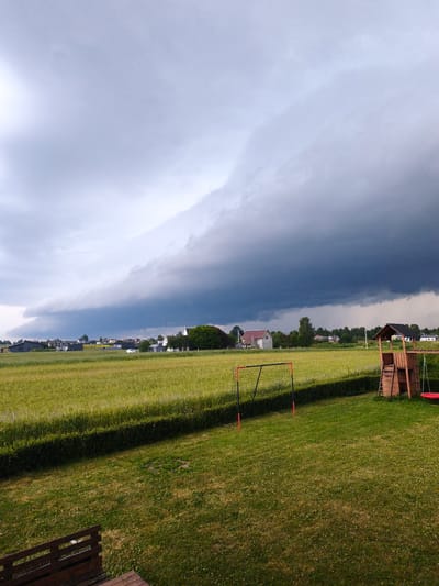 This is a medium shot of a rural scene under a dramatic, dark storm cloud formation. The foreground is a well-maintained green lawn with a low wooden railing visible in the bottom left corner. To the right, a wooden play structure with a slide and a swing is positioned on the grass. Further into the yard, a small soccer goal with a net stands ready for play.
Beyond the lawn, a vibrant green field stretches out, bordered by a dense, dark green hedge. In the middle ground, a village of houses with varying roof colors and architectural styles is nestled amongst trees. The sky above is dominated by a massive, dark, and ominous cloud bank that stretches across the frame, suggesting an approaching storm. The light is diffused and dim, indicative of pre-storm conditions.
The scene appears to be set in Dobromislė, Lithuania. The overall mood is one of impending weather change, with the beauty of the landscape contrasting with the power of the approaching storm. There are no visible people or text in the image. The time of day is unclear due to the overcast conditions, but it is likely daytime.
