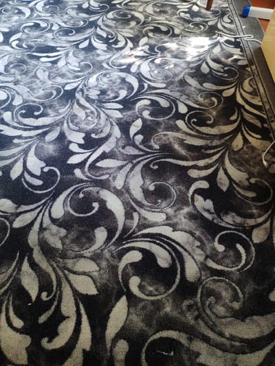 The image shows a close-up view of a dark grey carpet with a swirling, abstract floral pattern in a lighter grey and white. The carpet appears to be in an indoor setting, possibly a room or hallway. Sunlight is casting shadows and highlights across the surface of the carpet, indicating a window or light source is present. There are a few small specks of what appear to be dirt or debris scattered on the carpet. In the upper right portion of the image, a white electrical cord is visible, and a portion of a light blue and white electrical device is also partially visible. A dark brown wooden object, likely furniture, is in the far right background, and a dark brown edge of another rug is visible along the right side. The context provided by the prompt mentions the city of Первомайский, Russia, but this specific location cannot be determined from the visual information in the image alone.