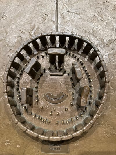 This image displays a bronze or brass tactile relief map of Stonehenge, located within the Stonehenge Visitor Centre near Amesbury, United Kingdom. The circular relief, set against a roughly textured metallic background, depicts the prehistoric monument's layout.
The outermost feature is a segmented ring of rectangular blocks, labeled "SARSEN CIRCLE" at the bottom of the relief. Inside this circle, several larger, three-dimensional upright stone structures are depicted, some forming trilithons with horizontal lintels. An inner arrangement of numerous smaller, peg-like protrusions represents the "BLUESTONES," labeled in a central, recessed oval plaque. Shadows are cast by the raised elements, indicating the three-dimensional nature of the relief. The entire piece has an aged, metallic sheen, consistent with bronze or brass.