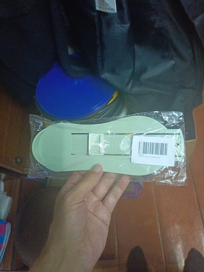 A light green shoe last is held in a person's hand, sealed in clear plastic packaging. The shoe last has a contoured shape designed to mimic the bottom of a shoe and features a white adjustable section with markings. A barcode label with Cyrillic text is affixed to the packaging. In the background, a blue circular object and a dark fabric are visible. The scene appears to be indoors, possibly in a room with wooden flooring and shelving, though its specific location in the city of Ukhany, Russia, cannot be determined from the image.