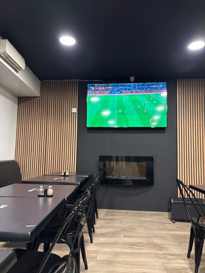 A modern interior features a black wall with two lights and a mounted TV showing a soccer game. A dark frame hangs near the TV. The opposite wall is adorned with horizontal wooden slats. Black tables and chairs line the right side of the frame, with flooring of light-colored wood planks. An air conditioning unit is positioned near the bottom-left corner.