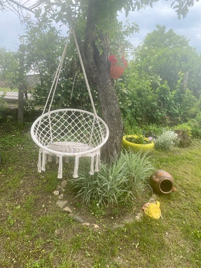 The image displays an outdoor garden scene in Kipilovo, Bulgaria, under an overcast sky. A white macrame swing chair, furnished with a circular beige cushion, is suspended from the trunk of a mature tree on the left side of the frame. An orange planter containing red flowers is affixed to the tree trunk above the swing. The ground beneath consists of green grass, scattered white petals, exposed soil, and a partial border of small stones. A plant with elongated, variegated green and white leaves grows in front of the swing. To the right of the tree, a yellow-painted tire serves as a planter, holding yellow and purple flowers. Further to the right, an earthenware pot and a yellow watering can lie overturned on the grass. The background is dominated by dense green foliage, partially obscuring a metal chain-link fence. No human or animal subjects are present within the frame.