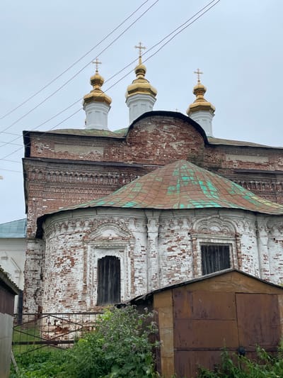 This image depicts the Trinity Church in the village of Kstinino, Russia, under an overcast sky. The church features a red brick exterior with white decorative elements, showing signs of age and weathering. Three golden onion domes with crosses are visible at the top, supported by white columns. The church has a green copper roof that is visibly oxidized. In the foreground, a rusty metal garage or shed stands, partially obscuring a metal fence and lush green vegetation. The overall impression is one of a historic religious building in a rural setting, showing the passage of time.