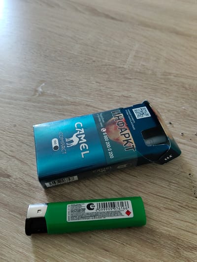 The image shows a close-up, eye-level view of a wooden surface, likely a table, with two smoking-related items on it. In the foreground, a green disposable lighter with black accents is positioned horizontally, with its nozzle facing left. The lighter displays text and a barcode, including "ЗАЖИГАЛКА DY-062" and a series of numbers. Behind the lighter and slightly to the right is a partially opened pack of Camel cigarettes. The pack is predominantly teal and black, featuring the iconic Camel logo of a dromedary camel. Text on the pack includes "CAMEL COMPACT," "100," and Russian text that translates to "INFARCT" and "More details about the consequences." A QR code is also visible on the pack. The lighting suggests it is daytime, and there are no obvious weather cues. The scene appears to be indoors, given the material of the surface. The items suggest a context related to smoking or preparing to smoke.