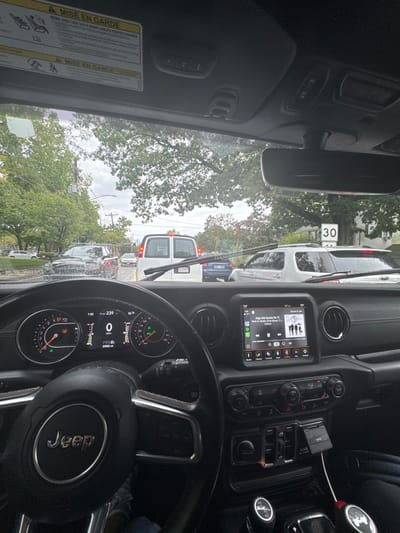 The image provides a driver's perspective from inside a Jeep Wrangler, stationary in a line of traffic on an overcast day. The vehicle's dashboard displays 0 MPH, 23082 miles, and 56°F. The infotainment system shows Apple CarPlay operating, playing "Algo Me Gusta De Ti" by Wisin & Yandel. A "MISE EN GARDE" warning label is affixed to the sun visor. Through the wet windshield, the immediate vehicle ahead is a white panel van with a Massachusetts license plate (3U5881). Behind it, a silver SUV and a dark blue sedan are visible in the queue. Green deciduous trees line both sides of the street, and a "30" MPH speed limit sign is posted on the right side of the road. The scene is located on a street in Newton, Massachusetts, United States.