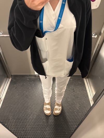 A person is standing in an elevator with a black zip-up jacket over a white uniform. A blue lanyard with white text is visible around the person's neck. A dark-colored smartphone is tucked into the left breast pocket of the white uniform, peeking out from under the jacket. The person is wearing white pants and beige sandals. The elevator floor is covered in a dark, textured mat. The elevator walls appear to be metallic. There is no discernible location within Harstad, Norway, that can be identified from this image.
