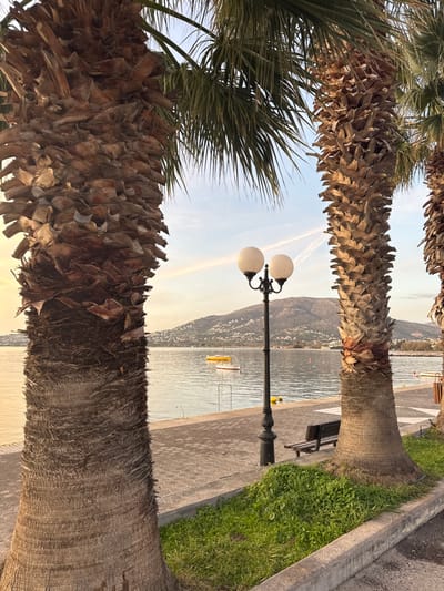 Palm trees frame a picturesque scene of a tranquil waterfront. A quaint bench rests along a paved path lined with grass. A classic street lamp with two globe lights stands tall. In the background, a calm sea reflects the soft light, with mountains visible across the water. A small yellow boat floats peacefully in the bay, adding to the serene atmosphere.