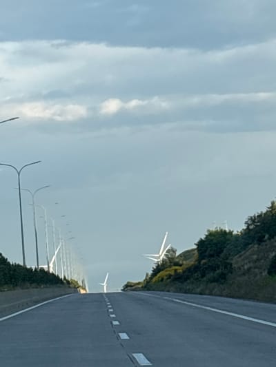 The image depicts a road in Otarsheni, Georgia, bordered by a line of streetlights on the left and a vegetated hillside on the right. In the distance, several wind turbines are visible, with one prominently featured on the right side of the road. The sky is overcast with clouds.