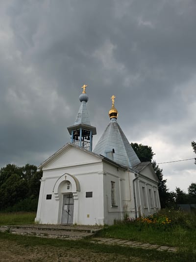 A white, Orthodox church with a bell tower and a gold dome stands against a dark, cloudy sky in Ulitkino, Russia. The church has a grey, pitched roof and a grey door with a cross above it. The bell tower has multiple bells and a golden cross on top. The church is surrounded by green grass, trees, and a few flowers. The ground in front of the church is paved with stone slabs.